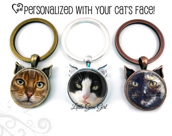 Op de afbeelding: Drie sleutelhangers met verschillende gekleurde metalen ringen en kattengezichtbedels. De bedels zijn rond met een glazen inzetstuk met een ander kattengezicht. De bedels zijn met een metalen lus aan de ringen bevestigd. De sleutelhangers zijn gepersonaliseerd met het gezicht van uw kat!