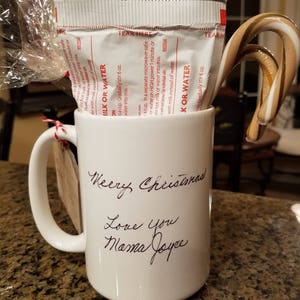 Może przedstawiać: Biały ceramiczny kubek z odręcznym napisem "Merry Christmas Love You Mama Joyce". Kubek zawiera paczkę mieszanki do gorącej czekolady i laska cukrowa. Do kubka przymocowana jest czerwona wstążka i etykieta.