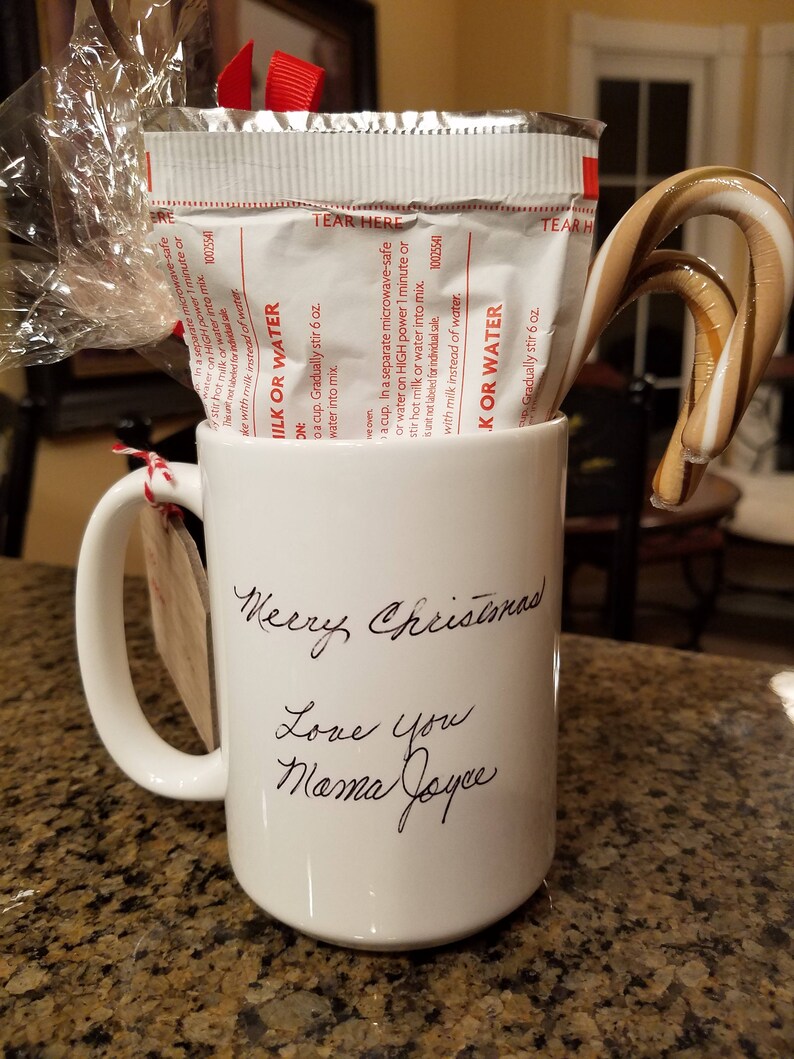 Może przedstawiać: Biały ceramiczny kubek z odręcznym napisem "Merry Christmas Love You Mama Joyce". Kubek zawiera paczkę mieszanki do gorącej czekolady i laska cukrowa. Do kubka przymocowana jest czerwona wstążka i etykieta.