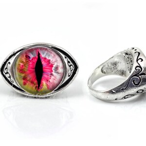 May include: A silver ring with an oval-shaped, pink, yellow, and black eye design. The ring has an intricate, silver, scroll-like design around the eye.