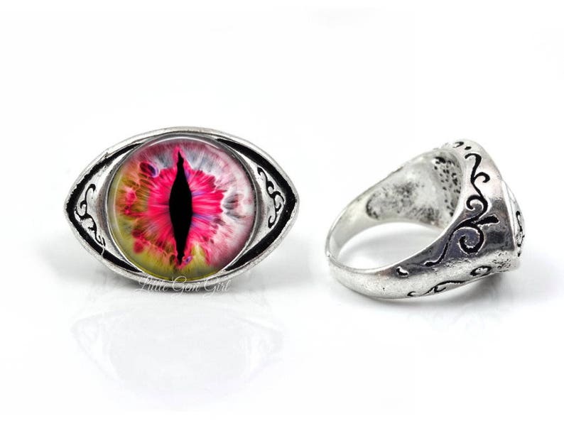 May include: A silver ring with an oval-shaped, pink, yellow, and black eye design. The ring has an intricate, silver, scroll-like design around the eye.