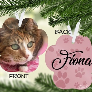 May include: Pink paw print shaped Christmas ornament with a cat photo on the front and the name "Fiona" on the back. The ornament is decorated with paw prints.