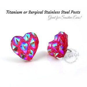 May include: A pair of heart-shaped earrings with a pink and iridescent finish. The earrings are made of surgical stainless steel or titanium and are great for sensitive ears. The text "Little Gem Girl" is visible in the image.