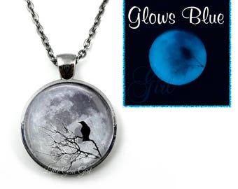 May include: A silver necklace with a round pendant featuring a black crow silhouette perched on a bare tree branch against a backdrop of a gray moon. The pendant glows blue in the dark.