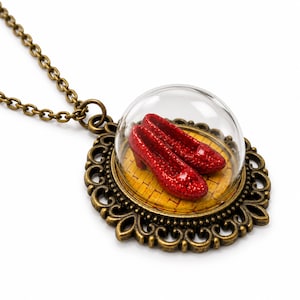 May include: A bronze-toned pendant necklace featuring a pair of miniature red glittery shoes encased under a clear glass dome. The shoes rest on a yellow brick-patterned base, surrounded by an ornate, filigree-style bronze metal frame.