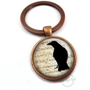May include: A bronze key chain with a round glass charm featuring a black silhouette of a raven on a vintage paper background. The text on the paper reads "While I nod as of someone's visit".