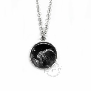 Puede incluir: Collar de plata con un colgante redondo que presenta una imagen de ultrasonido en blanco y negro. El colgante tiene el texto "Little Gem Girl Personalizado para Ti" alrededor del borde.