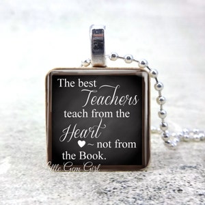 May include: A square pendant necklace with a silver-colored chain. The pendant has a chalkboard design with the text "The best Teachers teach from the Heart ~ not from the Book." The pendant is framed in a dark brown border.
