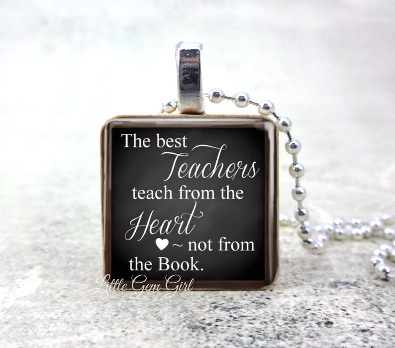 May include: A square pendant necklace with a silver-colored chain. The pendant has a chalkboard design with the text "The best Teachers teach from the Heart ~ not from the Book." The pendant is framed in a dark brown border.
