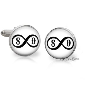 May include: A pair of silver cufflinks with white backgrounds and black infinity symbols with the letters "S" and "D" inside.