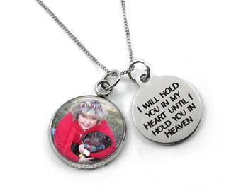 Op de afbeelding: Een zilveren ketting met een ronde fotobedel en een ronde bedel met de inscriptie "Ik zal je in mijn hart houden totdat ik je in de hemel vasthoud".