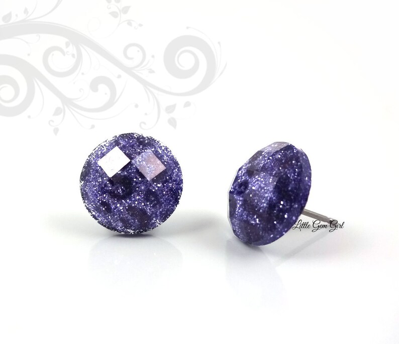 May include: A pair of purple glitter stud earrings. The earrings are round and have a faceted design. The glitter is a fine, sparkly texture.