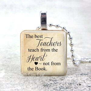 May include: A square pendant necklace with a silver-tone bail and ball chain. The pendant features the text "The best Teachers teach from the Heart ~ not from the Book." in black script on a beige background.