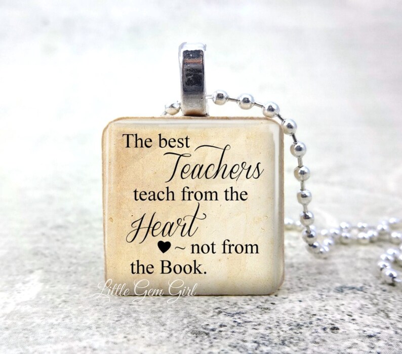 May include: A square pendant necklace with a silver-tone bail and ball chain. The pendant features the text "The best Teachers teach from the Heart ~ not from the Book." in black script on a beige background.