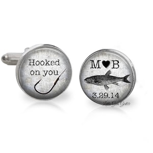 May include: A pair of silver cufflinks with a grey background. One cufflink features a fishing hook and the text "Hooked on you". The other cufflink features a fish, the initials "M & B", and the date "3.29.14".