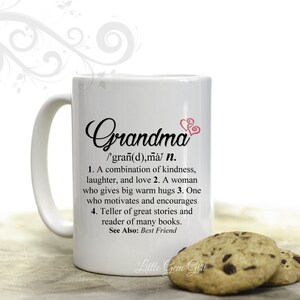 May include: White ceramic mug with a black and pink design. The mug features the word "Grandma" with a definition that reads: "/'gran(d),ma/ n. 1. A combination of kindness, laughter, and love 2. A woman who gives big warm hugs 3. One who motivates and encourages 4. Teller of great stories and reader of many books. See Also: Best Friend". The mug is sitting on a wooden surface with two chocolate chip cookies.
