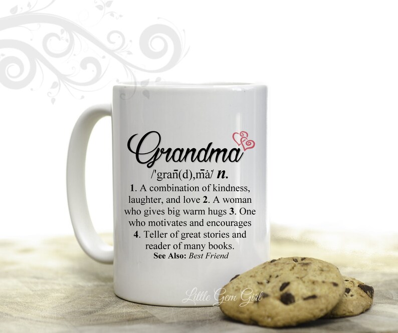 May include: White ceramic mug with a black and pink design. The mug features the word "Grandma" with a definition that reads: "/'gran(d),ma/ n. 1. A combination of kindness, laughter, and love 2. A woman who gives big warm hugs 3. One who motivates and encourages 4. Teller of great stories and reader of many books. See Also: Best Friend". The mug is sitting on a wooden surface with two chocolate chip cookies.
