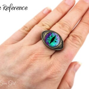 May include: A green and purple eye ring with a gold-toned setting. The ring is on a person's finger, showing the size of the ring.