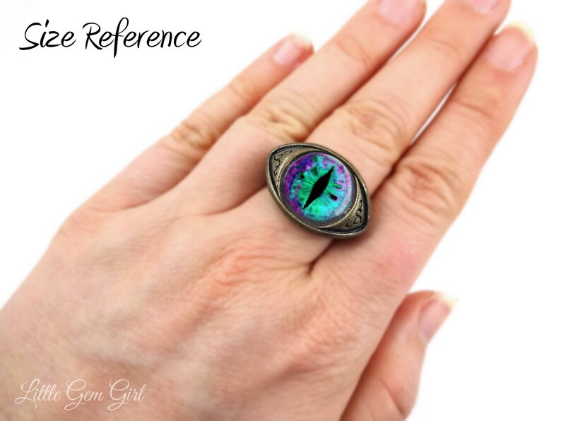 May include: A green and purple eye ring with a gold-toned setting. The ring is on a person's finger, showing the size of the ring.
