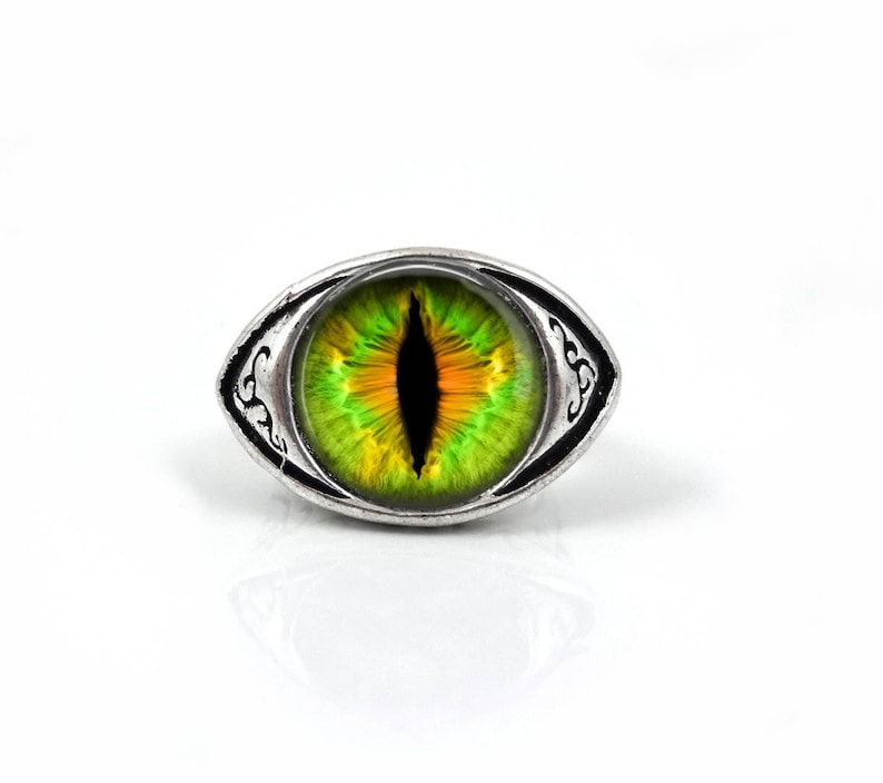 May include: A silver ring with a green and yellow eye design. The eye has a black pupil and a silver ring around it.
