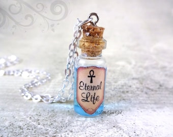 May include: A silver chain necklace with a small glass bottle pendant. The bottle is filled with a light blue liquid and has a cork stopper. A label on the bottle reads "Eternal Life" with an ankh symbol above the text. The label is cream colored with a brown border.