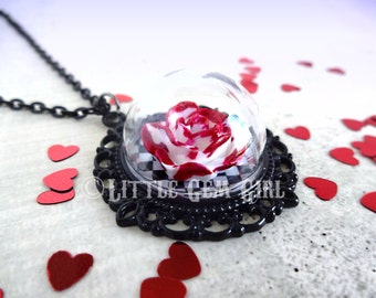 May include: A black necklace with a glass dome pendant containing a red and white rose on a black and white checkered background. The pendant is surrounded by a black filigree frame.