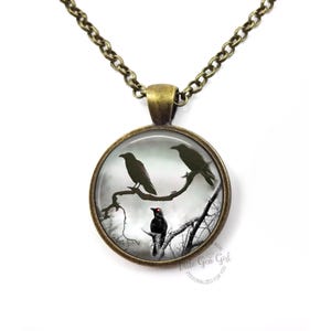 May include: A silver necklace with a round glass pendant featuring a black and white image of three crows perched on a bare tree branch. The crows are silhouetted against a cloudy sky.