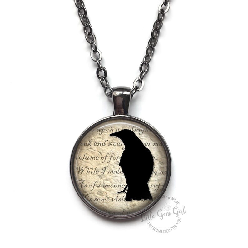 May include: A silver chain necklace with a round pendant featuring a black crow silhouette on a vintage-style paper background. The text on the paper reads "upon a...eak and wear...volume of for...While I nod...As of someone...some visit...erm...yna...rap".