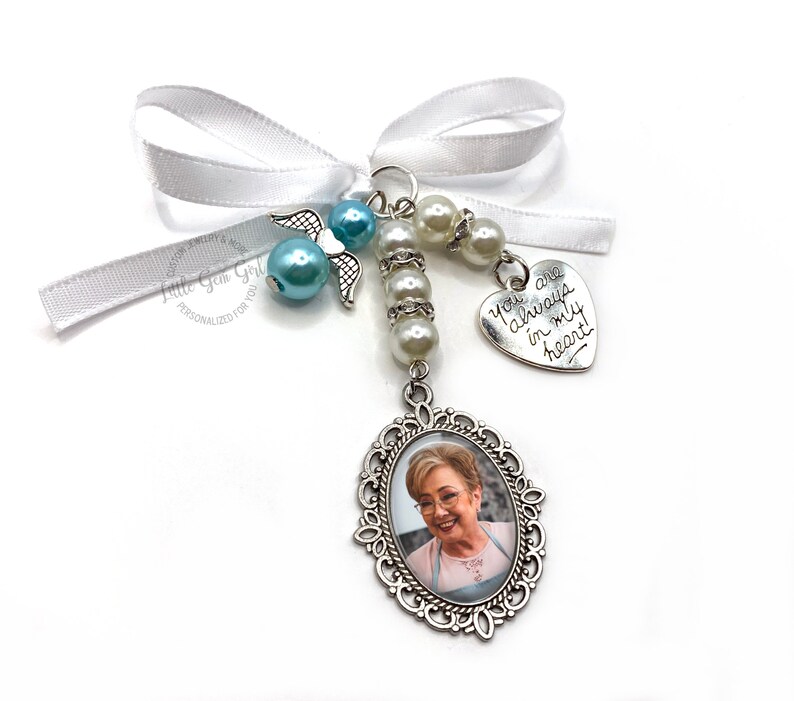 May include: A white satin ribbon with a silver charm featuring a photo of a woman. The charm is attached to a string of white pearls and a silver angel charm. The charm also has a silver heart with the inscription "You are always in my heart!"
