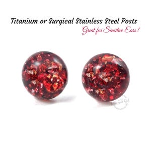May include: A pair of red stud earrings with gold flecks. The earrings are made of glass and have a round shape. The earrings are set on titanium or surgical stainless steel posts. "Great for Sensitive Ears!" is written in pink text on a white background.