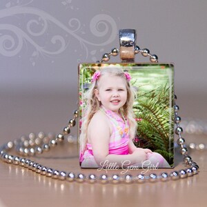 May include: A silver chain necklace with a square glass pendant featuring a photo of a young girl with blonde hair smiling. The pendant has the text "Little Gem Girl" printed on the bottom.