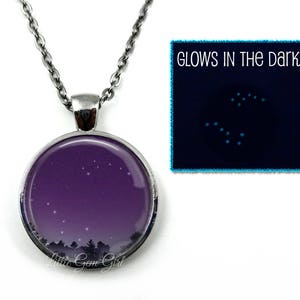 May include: A silver necklace with a round pendant featuring a purple night sky with white stars. The pendant glows in the dark, as shown in the inset image. "Glows in the Dark" is written in white text on a black background.