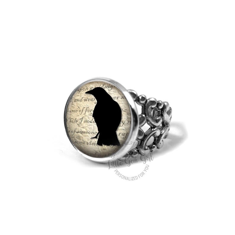 Pu&ograve; includere: Un anello d'argento con una silhouette di corvo nero su uno sfondo di pagina di libro vintage. L'anello &egrave; decorato con un design a filigrana decorativo. Il testo sulla pagina dice "and wear", "ver", "me of for", "hile I nede", "y", "of someone", "rup", "some visPRY", "Little Gem Girl", "PERSONALIZED FOR YOU".