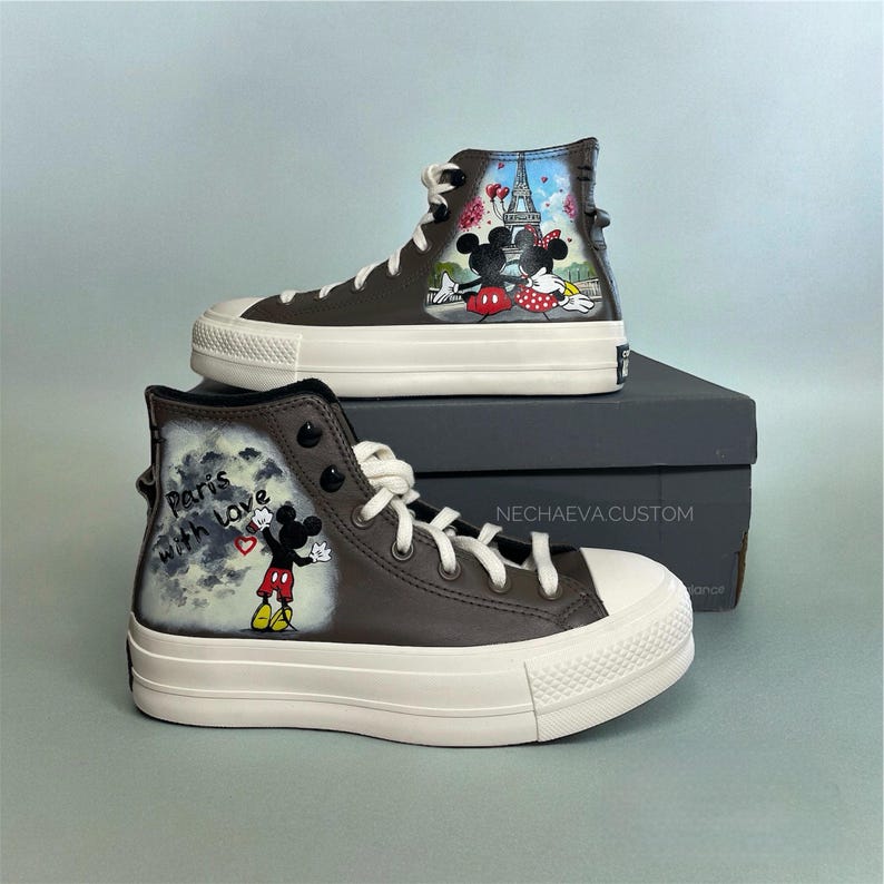 May include: Brown high-top sneakers with white soles and laces. The sneakers feature hand-painted artwork of Mickey and Minnie Mouse in Paris. One shoe has the text "Paris with love". The sneakers are on a gray box.