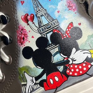 May include: Brown high-top sneaker featuring a hand-painted design of Mickey and Minnie Mouse in front of the Eiffel Tower. The artwork includes red heart balloons, pink flowers, and a blue sky background. The shoe has white laces and a white sole.
