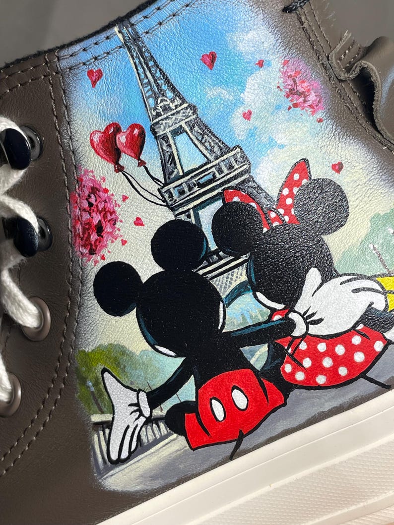 May include: Brown high-top sneaker featuring a hand-painted design of Mickey and Minnie Mouse in front of the Eiffel Tower. The artwork includes red heart balloons, pink flowers, and a blue sky background. The shoe has white laces and a white sole.