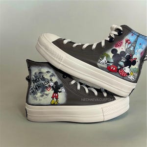 May include: Pair of custom high-top sneakers. The gray sneakers feature white laces and soles. One shoe has a painted scene of Mickey and Minnie Mouse in front of the Eiffel Tower. The other shoe says "Paris with love" with Mickey Mouse.