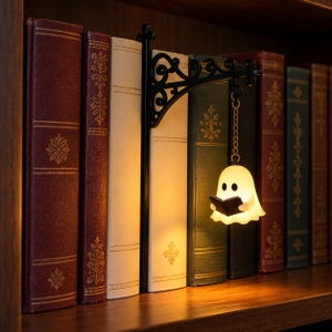 May include: A black metal bookend featuring an ornate, scroll-work bracket that extends outward. A small, glowing white ghost figurine holding a black book hangs from the bracket by a gold-toned chain, positioned between several hardcover books.