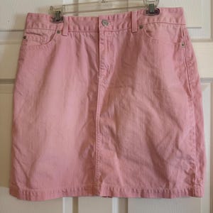 May include: A light pink denim skirt with a classic five-pocket design. The skirt has a button and zipper closure, belt loops, and a straight hemline. The skirt is hanging on a clear plastic hanger.