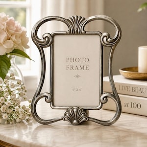 May include: Ornate silver-toned photo frame with a decorative shell and scroll design. The frame holds a 4" x 6" photo and features the words "PHOTO FRAME". The frame is displayed on a marble surface.