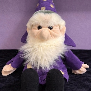 May include: A plush wizard doll with a purple robe and hat, featuring gold polka dots. The doll has a long white beard, a pink nose, and black shoes. The doll is sitting on a dark blue surface.