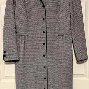 May include: A long, button-down coat in a black and white houndstooth pattern. The coat has a notched collar and black trim along the edges, cuffs, and button line. The coat is hanging on a black hanger.