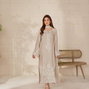 K&ouml;nnte beinhalten: Ein hellbeige, lang&auml;rmeliges Kleid mit aufwendiger wei&szlig;er Stickerei und Spiegelverzierungen. Das Kleid hat einen passenden Schal und wird mit goldenen Sandalen kombiniert. Geeignet f&uuml;r besondere Anl&auml;sse.