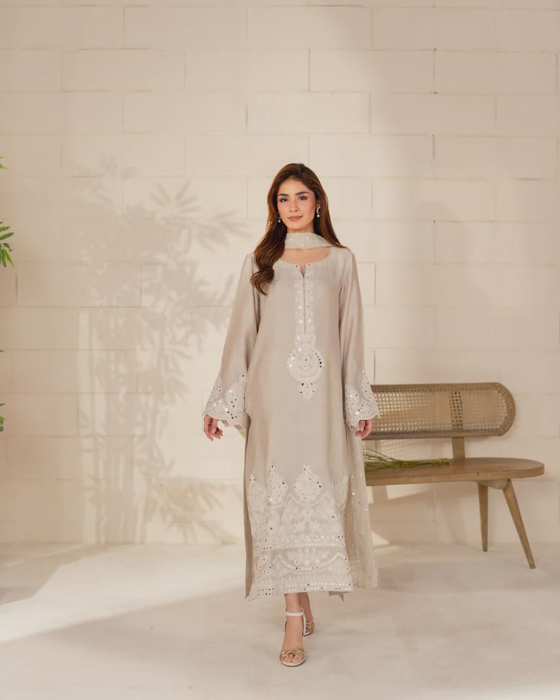 K&ouml;nnte beinhalten: Ein hellbeige, lang&auml;rmeliges Kleid mit aufwendiger wei&szlig;er Stickerei und Spiegelverzierungen. Das Kleid hat einen passenden Schal und wird mit goldenen Sandalen kombiniert. Geeignet f&uuml;r besondere Anl&auml;sse.
