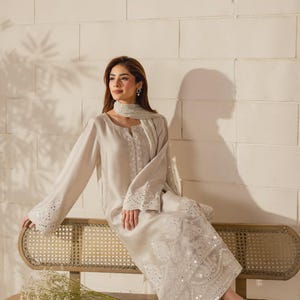 K&ouml;nnte beinhalten: Hellbeige Kleid mit langen &Auml;rmeln, verziert mit wei&szlig;en Stickereien und Pailletten. Das Kleid hat ausgestellte &Auml;rmel und einen passenden Schal. Das Model tr&auml;gt goldene und silberne Sandalen. Geeignet f&uuml;r besondere Anl&auml;sse.