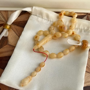 Puede incluir: Un collar de cuentas de oración de color crema con cuentas ovaladas y una borla, sobre una bolsa de satén blanco. Las cuentas están conectadas por un hilo rojo. La bolsa tiene un cierre de cordón con detalles dorados.