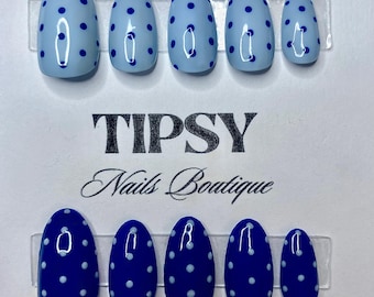 Pode incluir: Unhas postiças ovais em dois conjuntos. Um conjunto é azul claro com bolinhas azuis escuras, e o outro é azul escuro com bolinhas brancas. O texto "TIPSY Nails Boutique" está impresso no cartão.