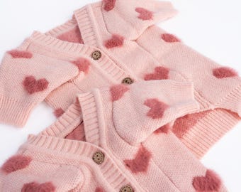 May include: Two pink knit cardigans with fuzzy heart appliqués. Each cardigan features a button-up front and ribbed cuffs. The hearts are a darker shade of pink, creating a contrasting design. The cardigans are displayed on a white background.