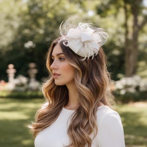 May include: A white headpiece with a bow and feathers, accented with netting. The headpiece is worn on the head of a person with long, wavy, light brown hair. The person is wearing a white dress, and the background is a blurred outdoor scene.