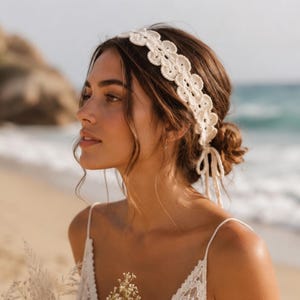 May include: A cream-colored crochet headband with a scalloped design. The headband is worn by a person with brown hair styled in a low bun. The person is wearing a white lace top, with a beach and ocean in the background.